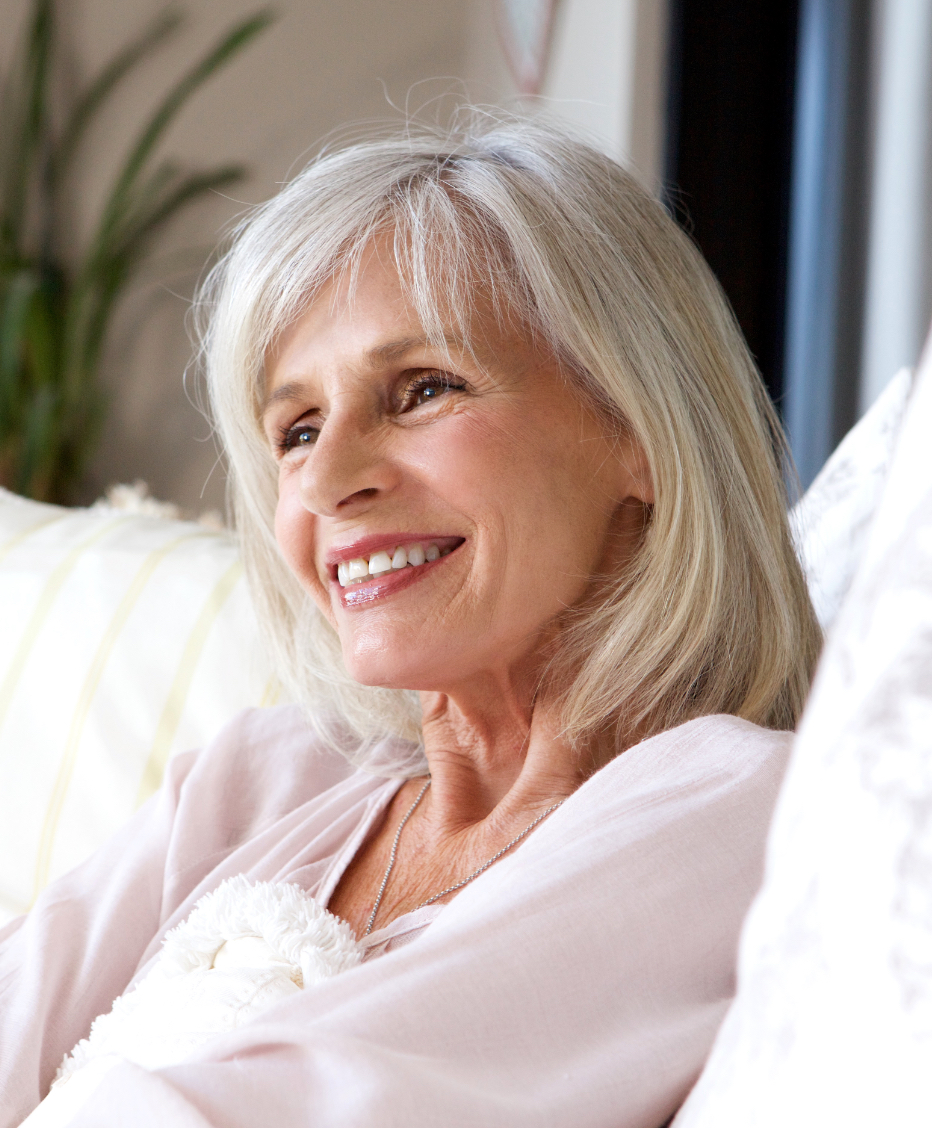 Femme dans la soixantaine aux cheveux blancs, souriant de profil à la caméra sur son canapé de velours blanc crème.