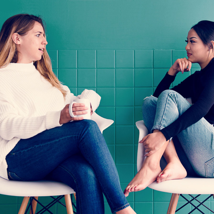 Deux jeunes femmes dans la vingtaine assises sur des chaises blanches, et discutant devant un mur turquoise.