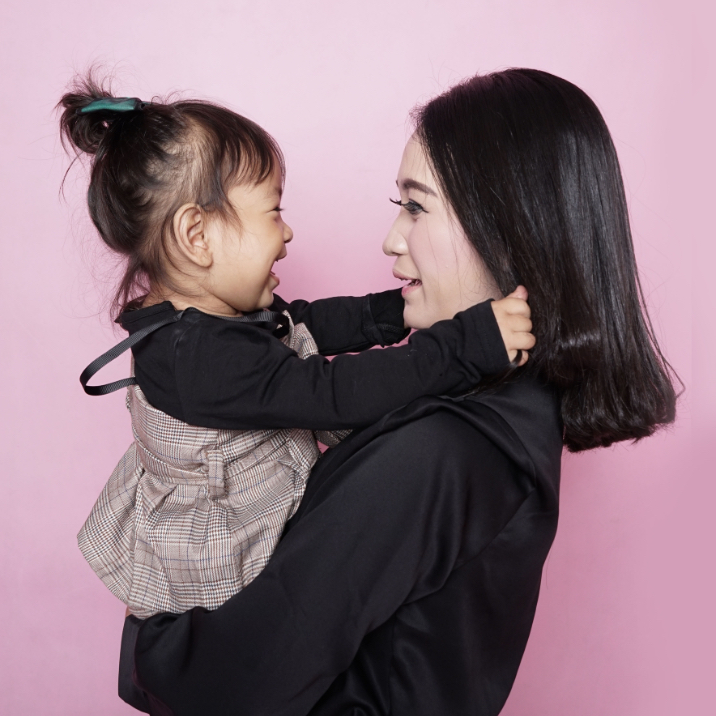 Jeune maman asiatique aux cheveux foncés portant dans ses bras sa petite fille âgée d'environ 3 ans, qui lui fait un beau sourire resplandissant.