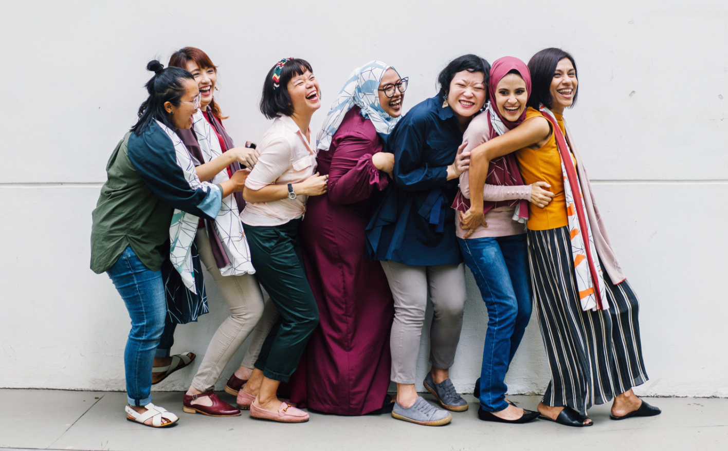 7 femmes de plusieurs ethnies s'accolant devant une mur gris pâle, souriant à pleines dents.