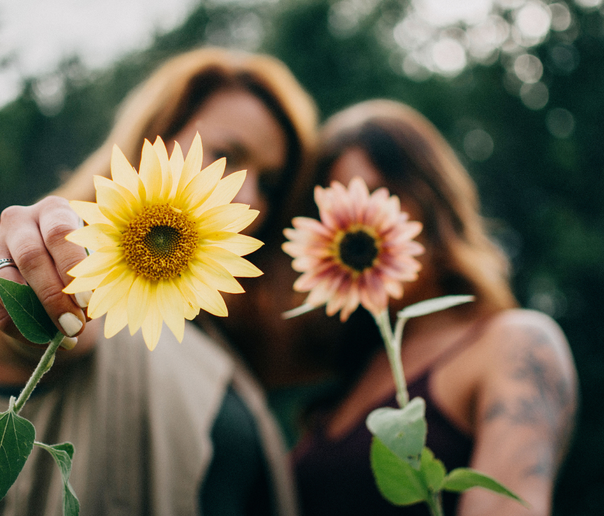 Deux jeunes femmes se donnant un câlin, tenant dans leurs mains des fleurs (tournesols) roses et jaunes. L'une des femmes est tattouée et l'autre porte des bagues aux doigts.